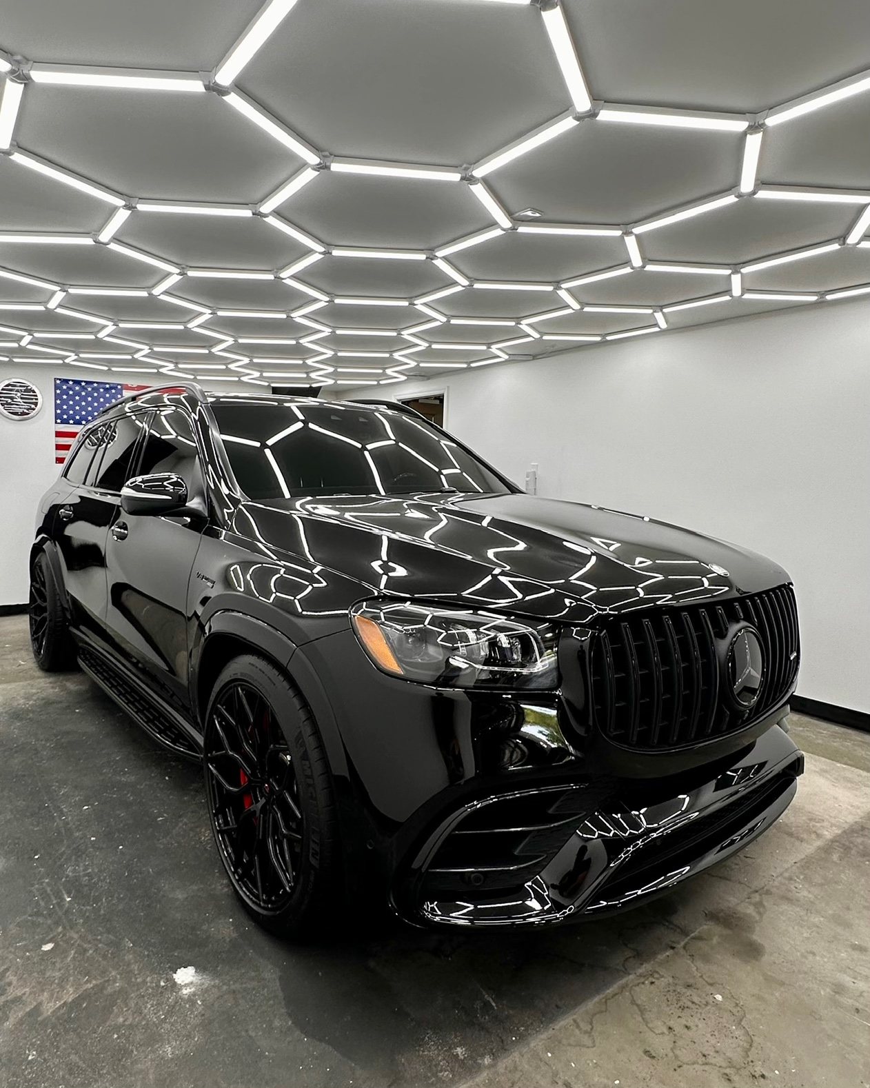 A black Mercedes-Benz GLS with a mirror-like corrected finish reflecting the garage's hexagonal lights